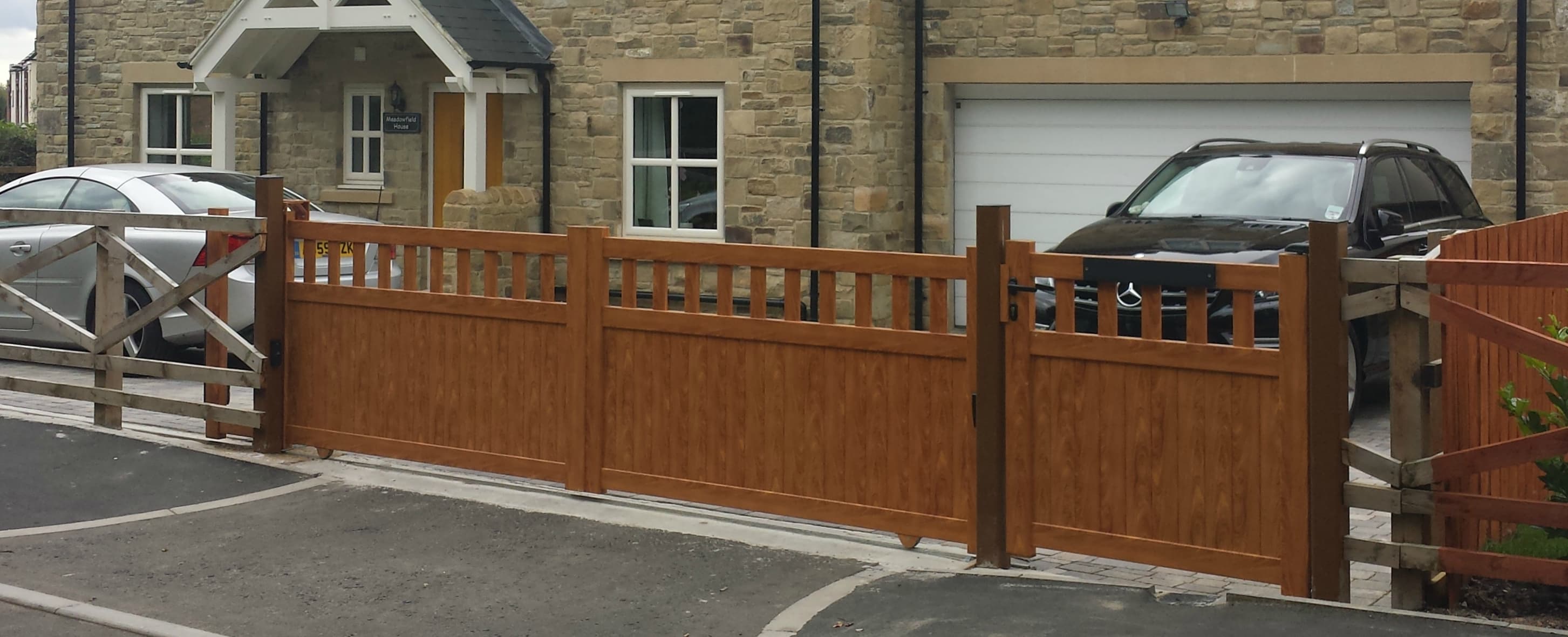 Aluminium driveway gates with a wooden appearance in front of a stone house with parked cars.